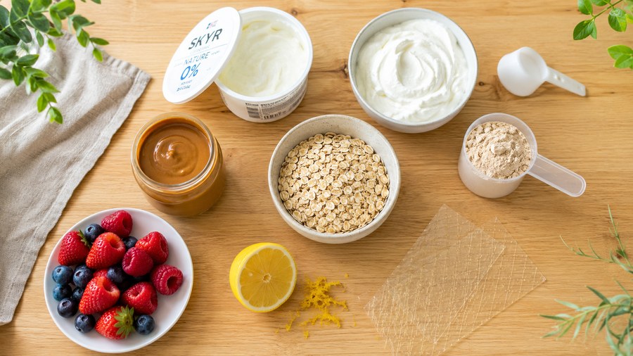 Ingrédients du cheesecake protéiné sans cuisson disposés à plat : skyr, fromage blanc, whey vanille, flocons d'avoine, purée de cacahuète, fruits rouges, citron, gélatine